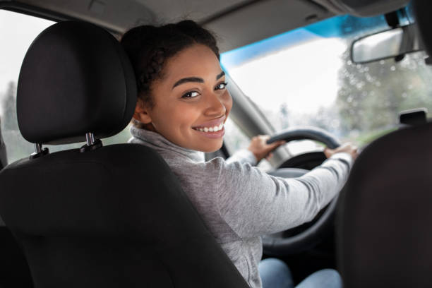 Cute driver, auto blogger and traveling with friends. Smiling beautiful young african american female turns to back seat and talk with passenger or client, view of window with raindrops, copy space